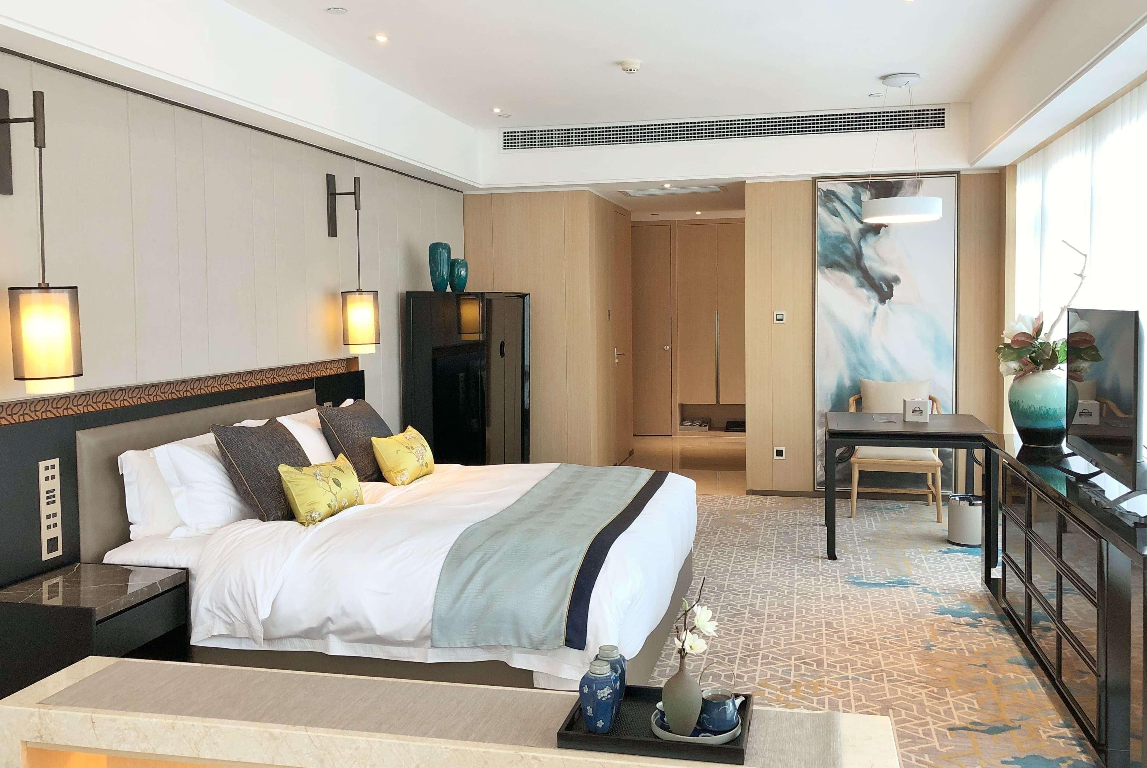 A hotel room with a bed, bedside tables, a desk, a chair, a wardrobe, a painting, and a vase.