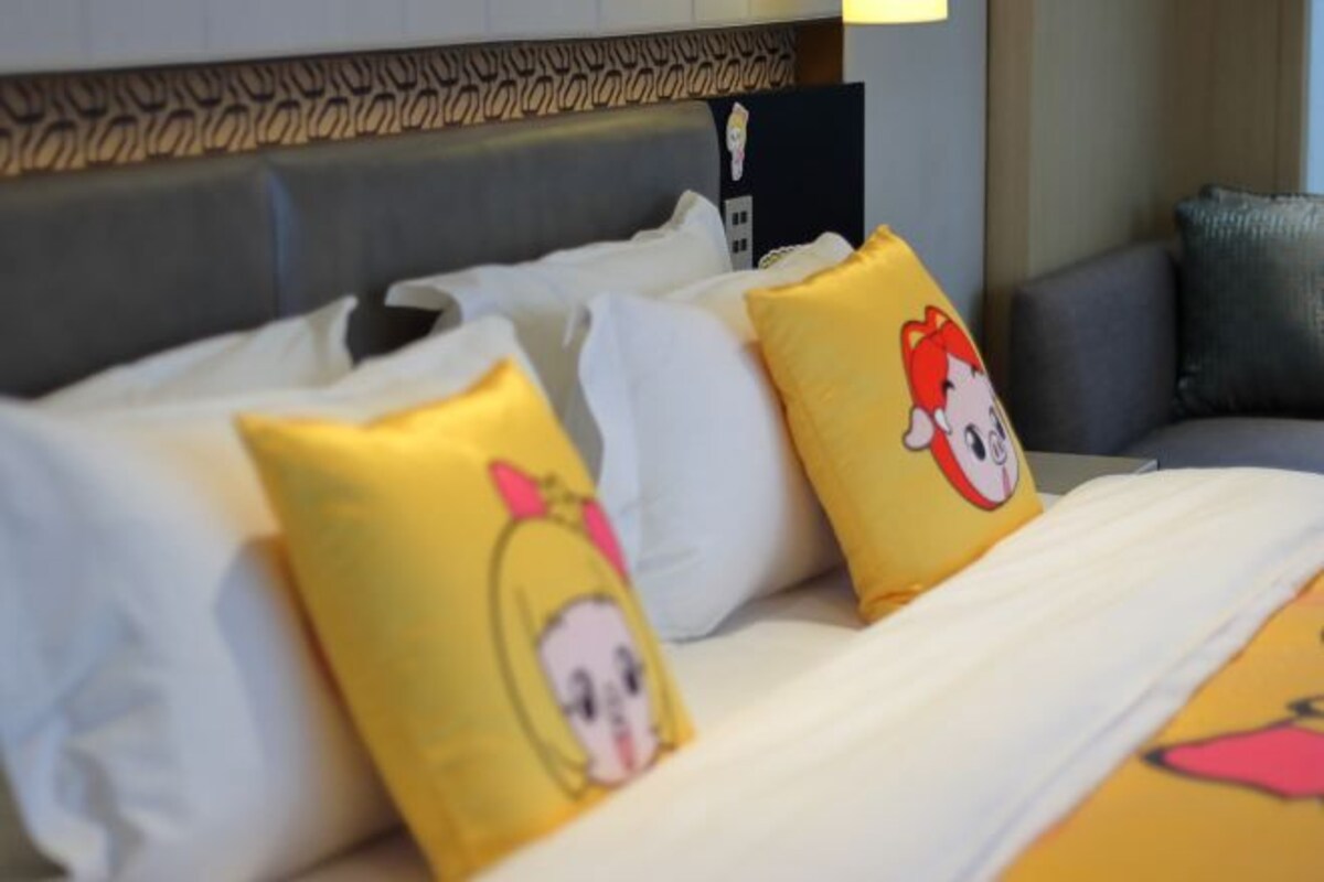 Une chambre d’hôtel avec un lit agrémenté d’oreillers blancs et jaunes ornés de personnages de dessins animés, une tête de lit grise et un canapé gris en arrière-plan.