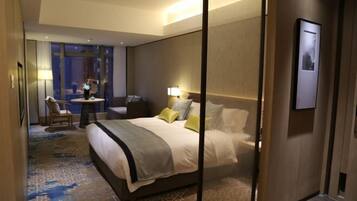 Habitación de hotel con una cama grande, un escritorio, una silla y vistas a la ciudad a través de amplios ventanales.