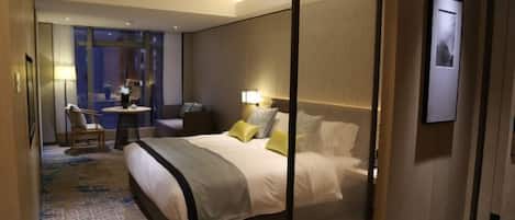 A hotel room with a large bed, a desk, a chair, and a view of the city through large windows.
