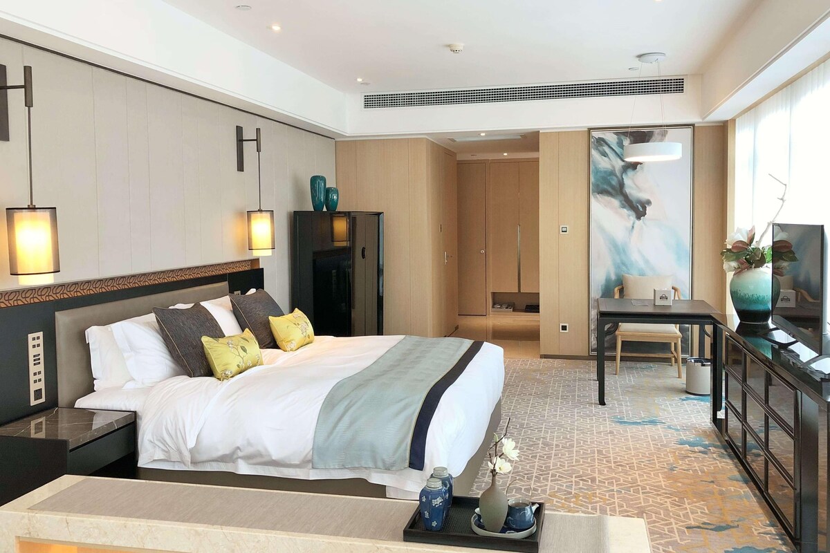 Une chambre d’hôtel comprenant un lit, des tables de chevet, un bureau, une chaise, une armoire, un tableau et un vase.