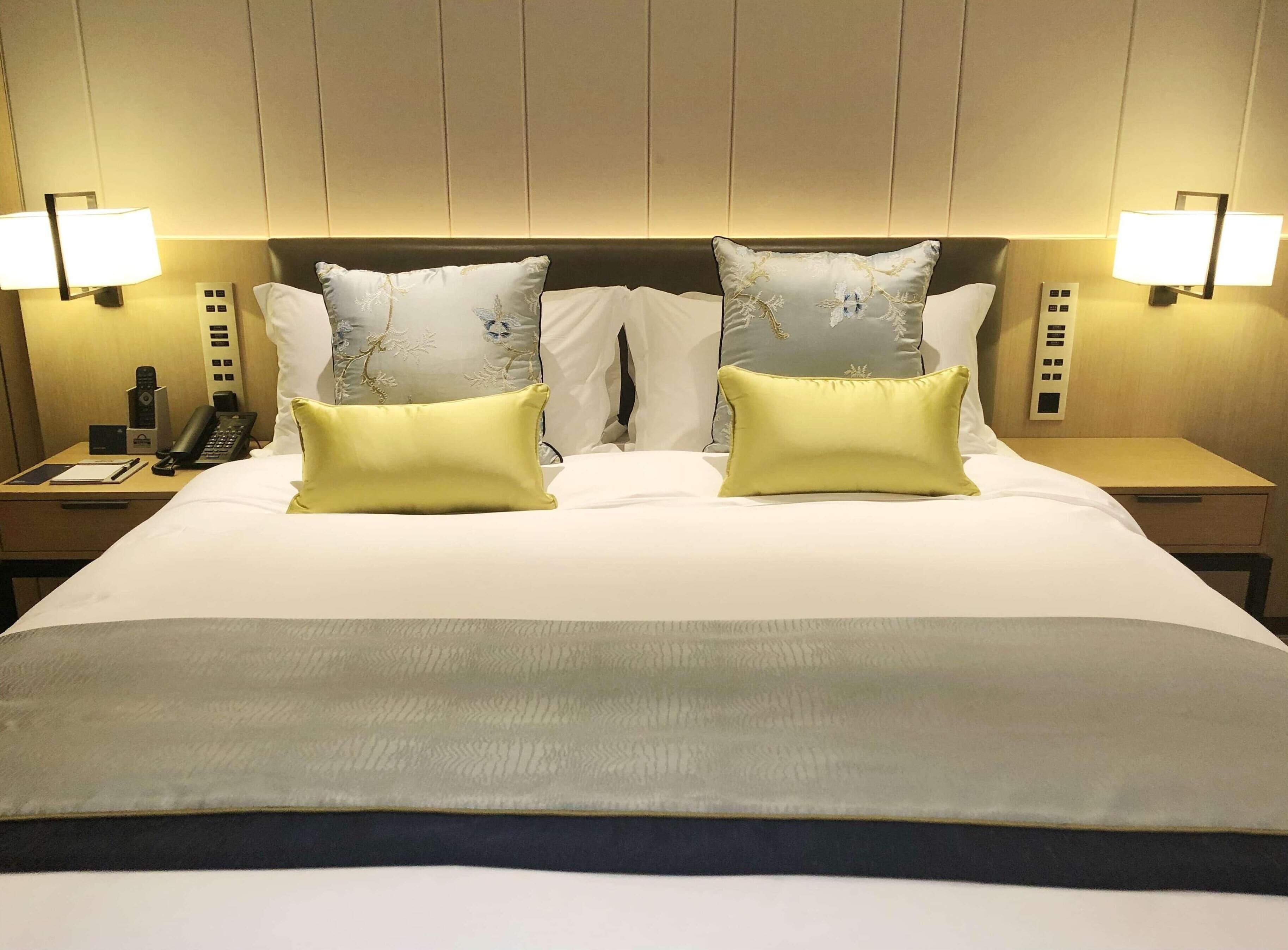 A hotel room with a large bed, two bedside lamps, a nightstand with a phone, and a wall-mounted control panel.