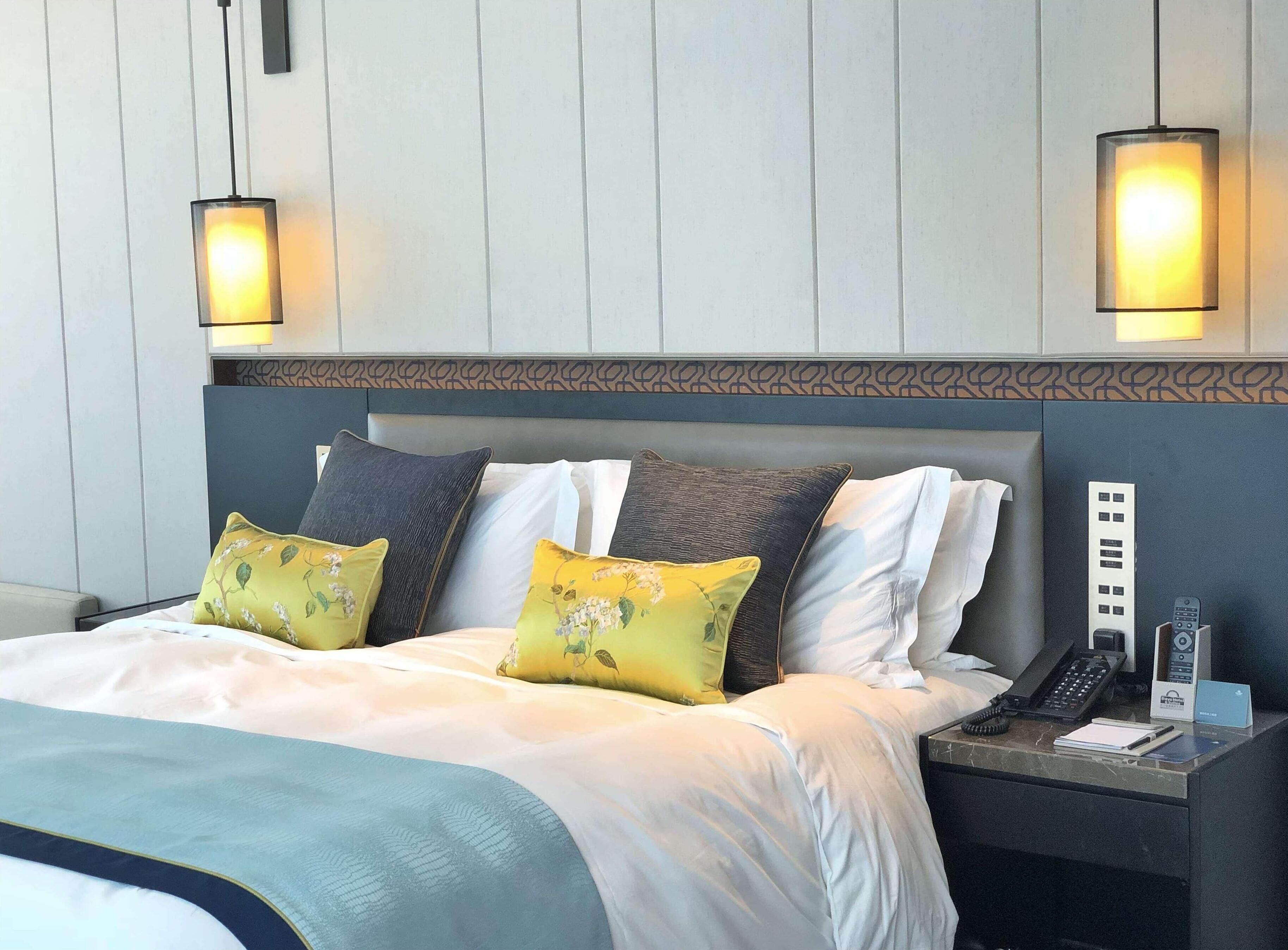 A hotel room with a bed, two bedside lamps, a nightstand with a phone, and a wall-mounted control panel.