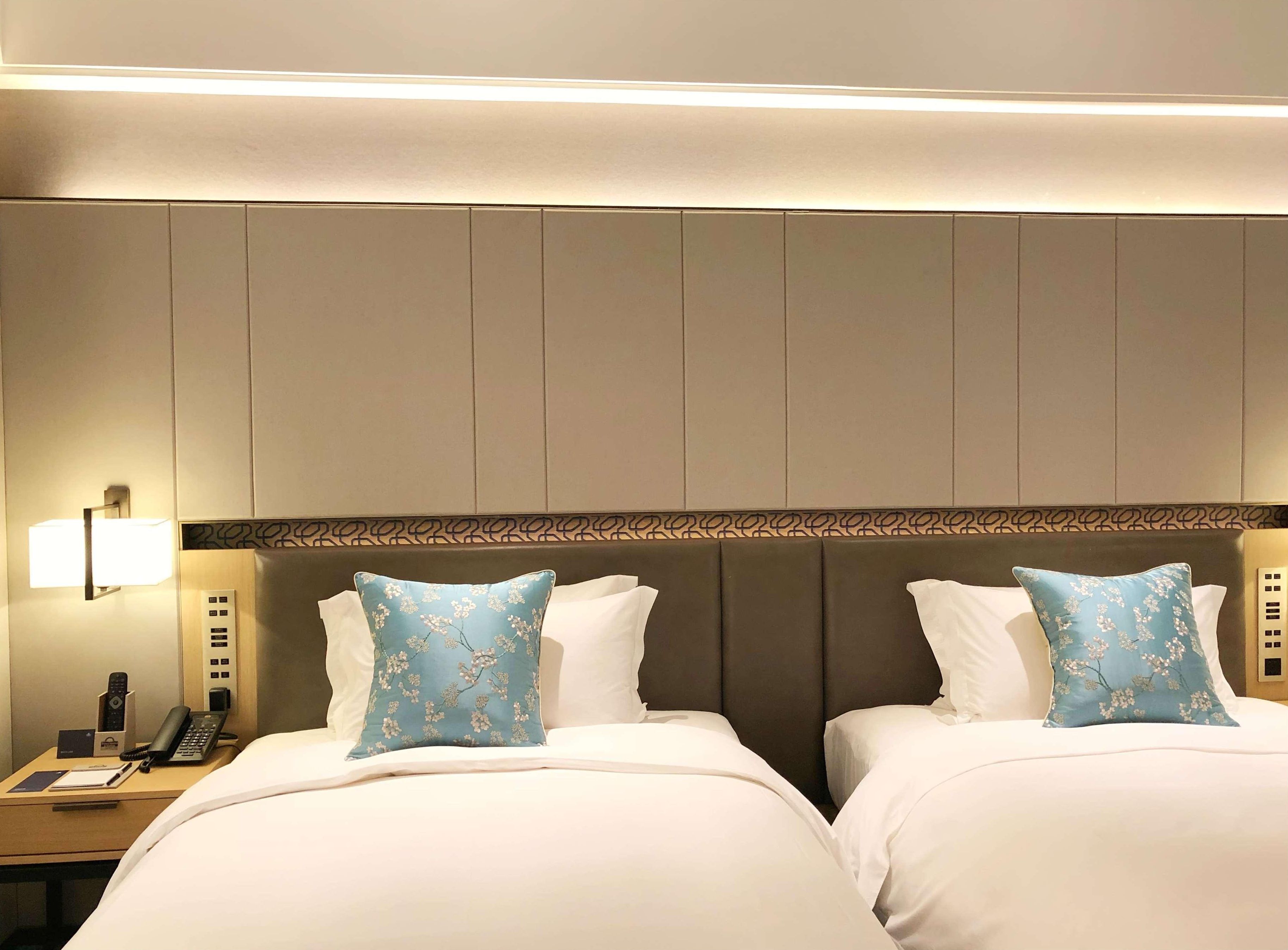 A hotel room with two beds, a bedside table with a phone and a lamp, and a patterned headboard.