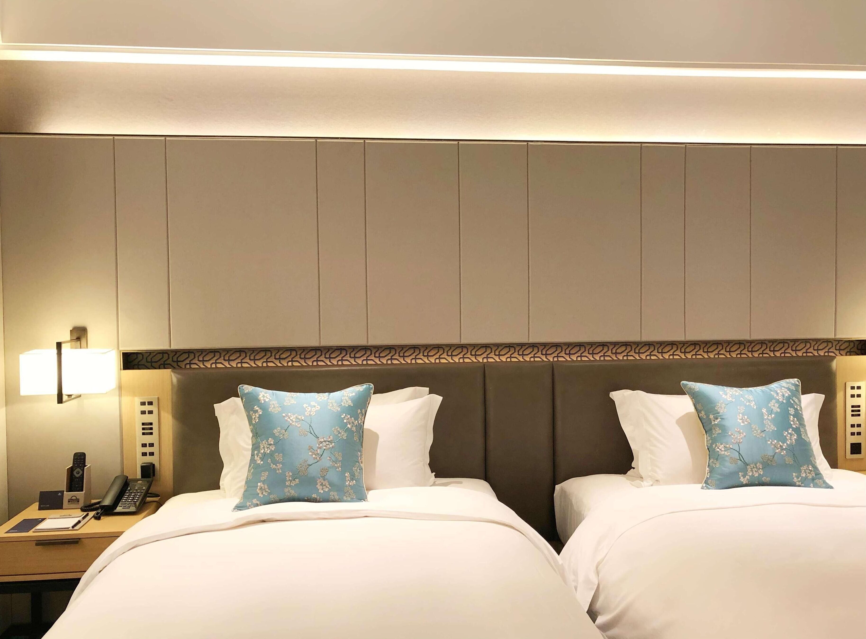 A hotel room with two beds, a bedside table with a phone and a lamp, and a patterned headboard.