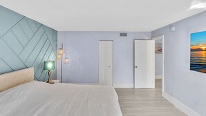A bedroom with a bed, a nightstand, a lamp, a wall-mounted light fixture, a door, and a painting of a sunset. - One Bedroom Steps Away From The Beach | Pool! (Sunny Isles Beach)