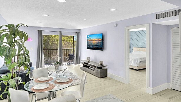 A modern living room with a glass dining table, a comfortable sofa, and a large window with a view of trees. - One Bedroom Steps Away From The Beach | Pool! (Sunny Isles Beach)