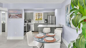 A modern kitchen with grey cabinets, stainless steel appliances, and a dining area with a glass table and white chairs. - One Bedroom Steps Away From The Beach | Pool! (Sunny Isles Beach)