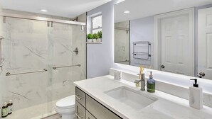 A modern bathroom with a glass-enclosed shower, a white sink with a large mirror, and a toilet. - One Bedroom Steps Away From The Beach | Pool! (Sunny Isles Beach)