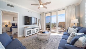A spacious living room with a large flat-screen TV, a ceiling fan, and a balcony with a view. - Azure 118 ~ 4BR Beachfront Ground Floor Corner Condo ~ Sleeps 14 ~ Must See! (Fort Walton Beach)
