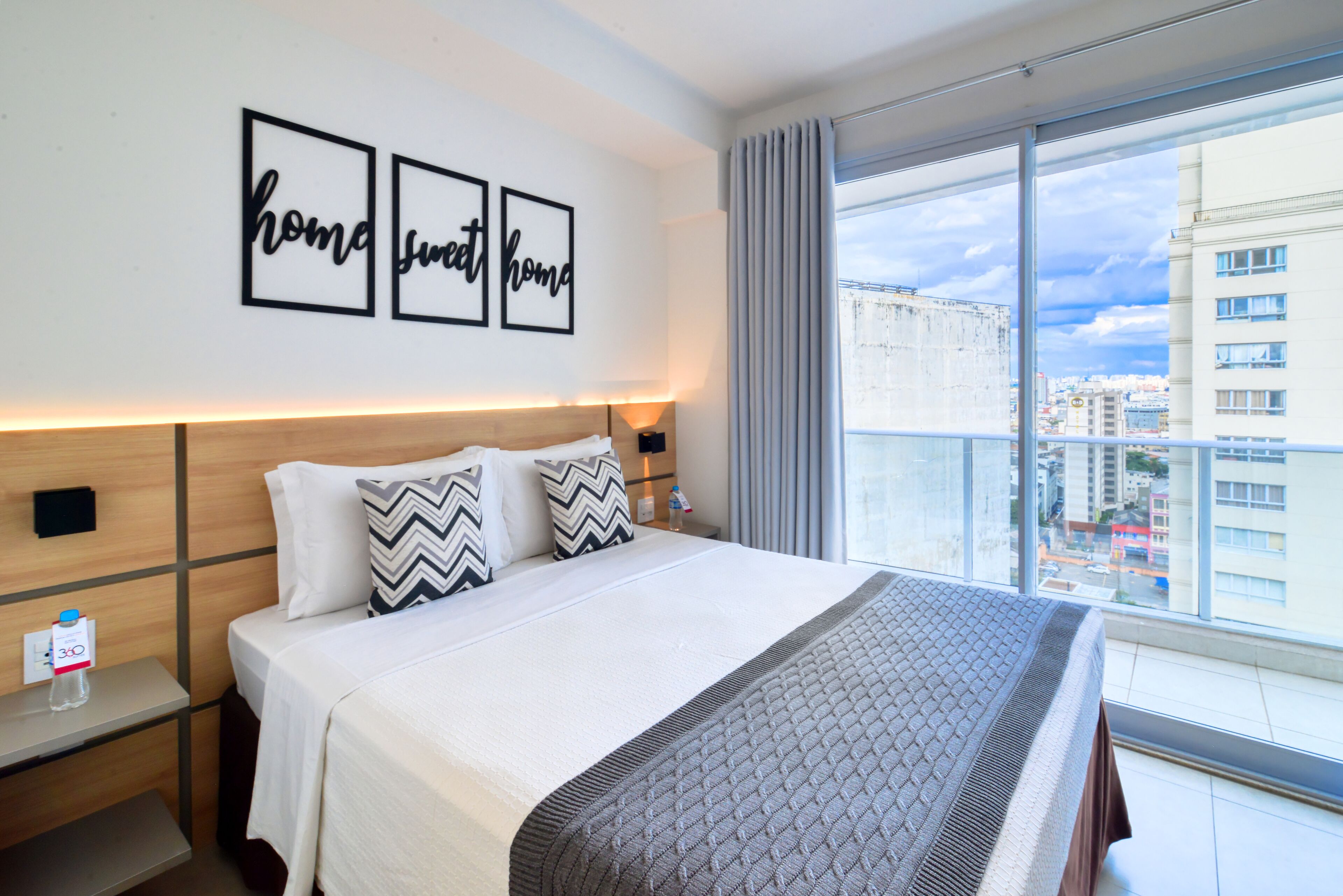 A hotel room with a large bed, a nightstand, a wall-mounted lamp, and a window with a city view.