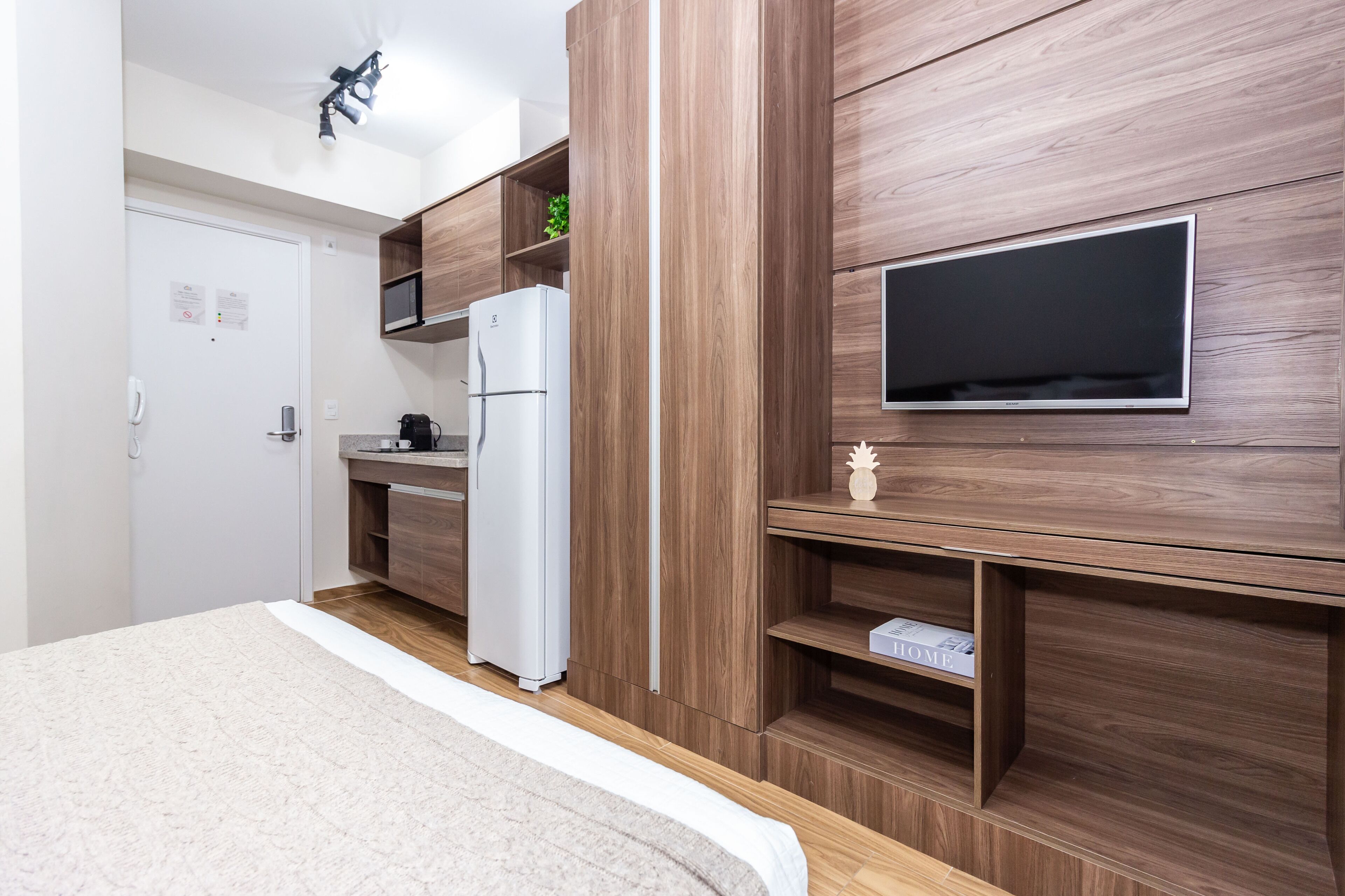 Una habitación de hotel moderna con cama, televisor de pantalla plana montado en la pared, refrigerador integrado y una pequeña cocina con microondas y cafetera.