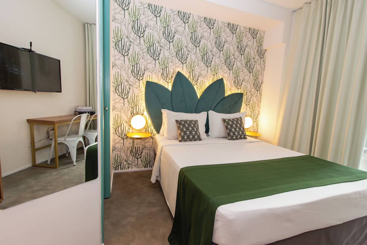 A hotel room with a bed, a desk, and a chair. The room has a cactus-themed wallpaper and a flat-screen TV mounted on the wall.