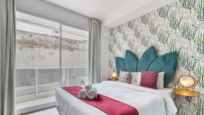 Habitación de hotel con un ventanal grande, cama con colcha rojo y blanco, almohadas color turquesa y papel tapiz con motivos de cactus.
