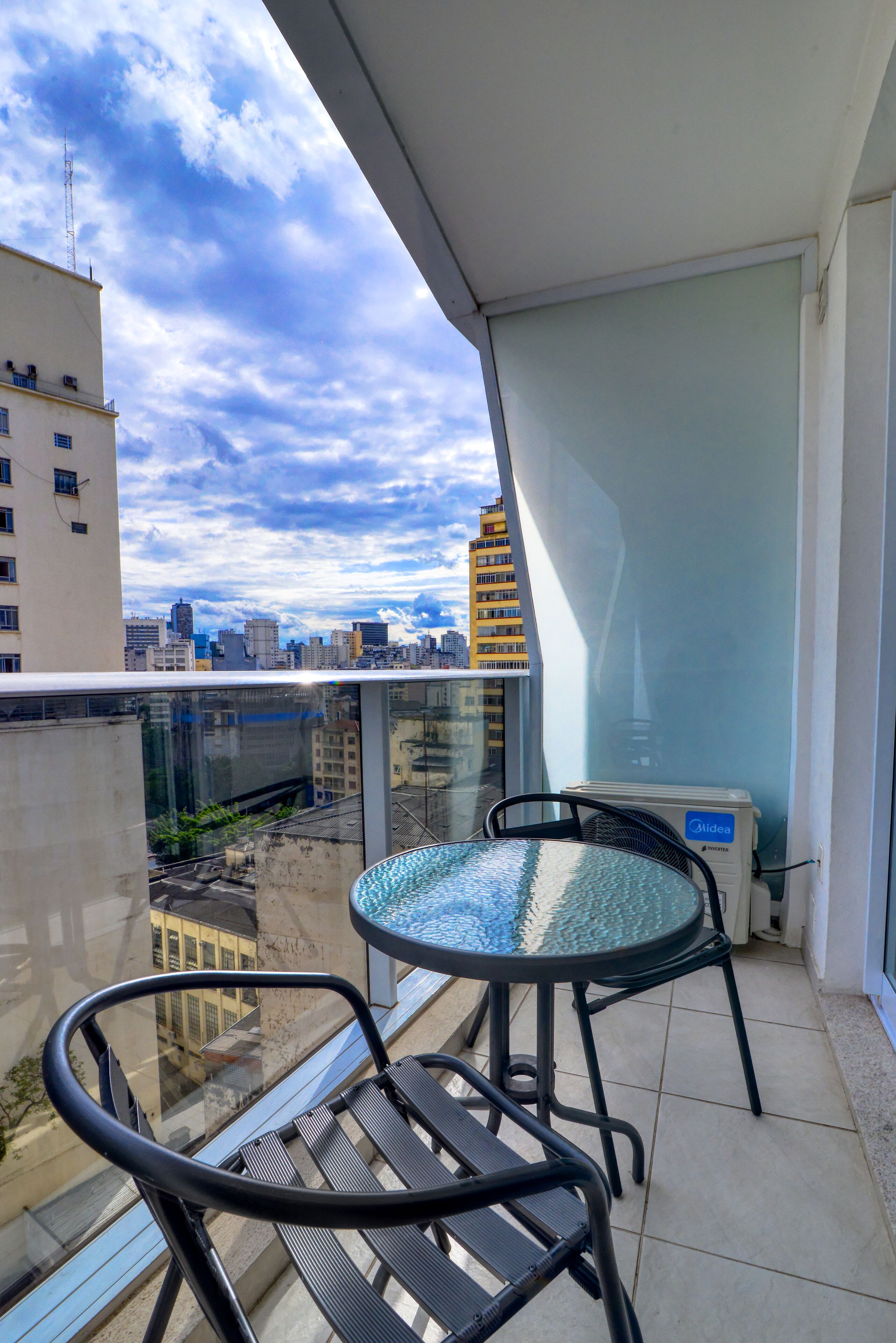 Un balcón con una mesa y sillas de vidrio, con vistas a un paisaje urbano con edificios y un cielo parcialmente nublado.