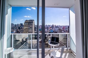 Balcón con una laptop, una silla y una mesa, con vista a un paisaje urbano lleno de edificios.