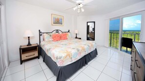 A bedroom with a bed, two bedside tables, a ceiling fan, a large window with a view, and a painting on the wall.