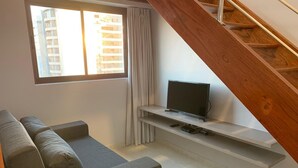 A modern living room with a grey sofa, a flat-screen TV on a white shelf, a window with curtains, and a wooden staircase. - Excelentes Duplex Maceió (Maceió)