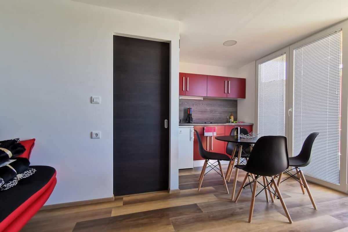 A compact living space with a kitchenette, a dining area with modern chairs, and a large window with blinds.