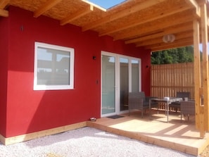 A covered patio with a wooden ceiling, a red wall, and a sliding glass door leading to an outdoor seating area with a table and chairs. - Village Lipa Ljubljana (Medvode)