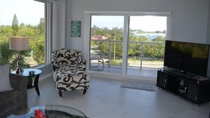 A spacious living room with a large window, a patterned armchair, a glass coffee table, a flat-screen TV, and a balcony with a view of greenery and buildings. - Beachcomber unit 507 (Longboat Key)