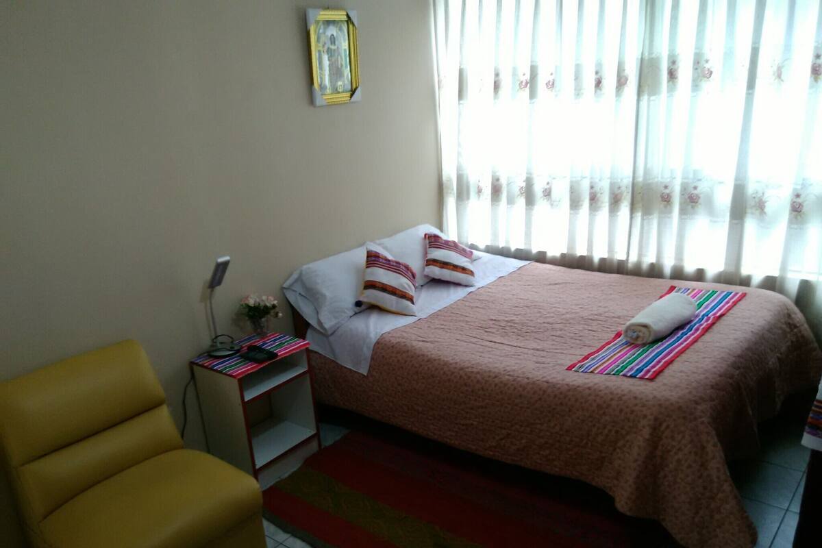 A hotel room with a bed, a yellow armchair, a bedside table with a lamp, and a window with floral curtains.
