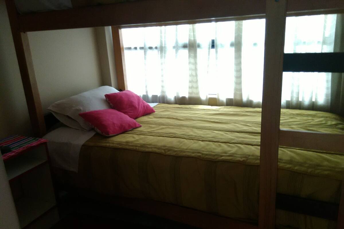 A bunk bed with a yellow mattress and pink pillows, a white pillow, and a small shelf with books.