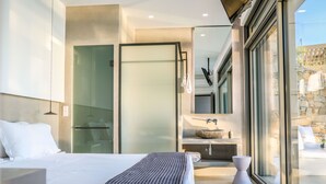 Una habitación de hotel moderna con una cama grande, cabina de ducha de vidrio, baño con lavamanos de piedra y vistas al exterior.