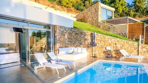 Un área de piscina moderna al aire libre con sillones de descanso, un muro de piedra y una sala con paredes de vidrio.
