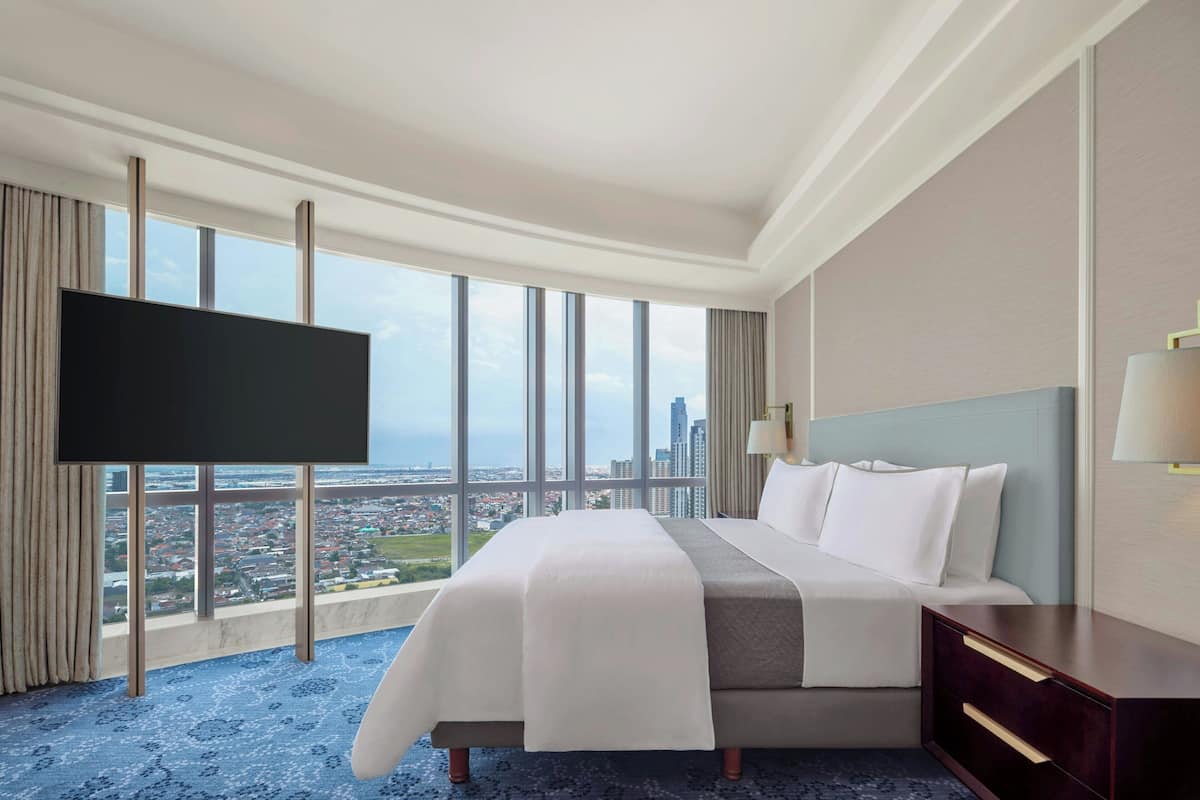 Habitación de hotel con una cama grande, televisión de pantalla plana, mesita de noche y vistas a un paisaje urbano a través de amplias ventanas.