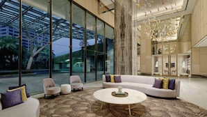 Een moderne lobby met een grote glazen wand, een centrale zithoek met een bank en fauteuils, en een decoratief element aan het plafond.