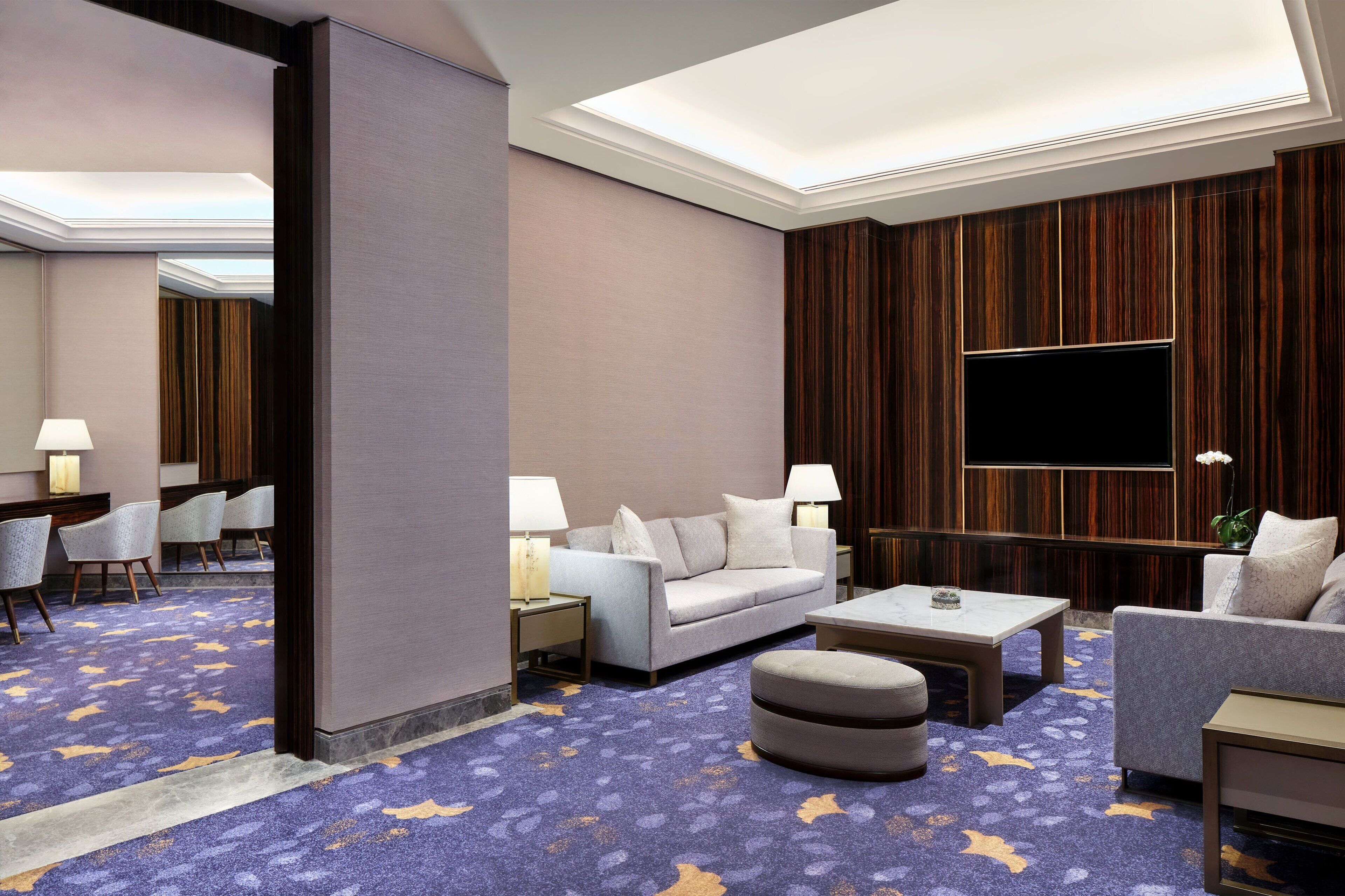 Une chambre d’hôtel dotée d’un grand téléviseur à écran plat, d’un canapé gris, d’un pouf rond et d’un tapis à motifs.