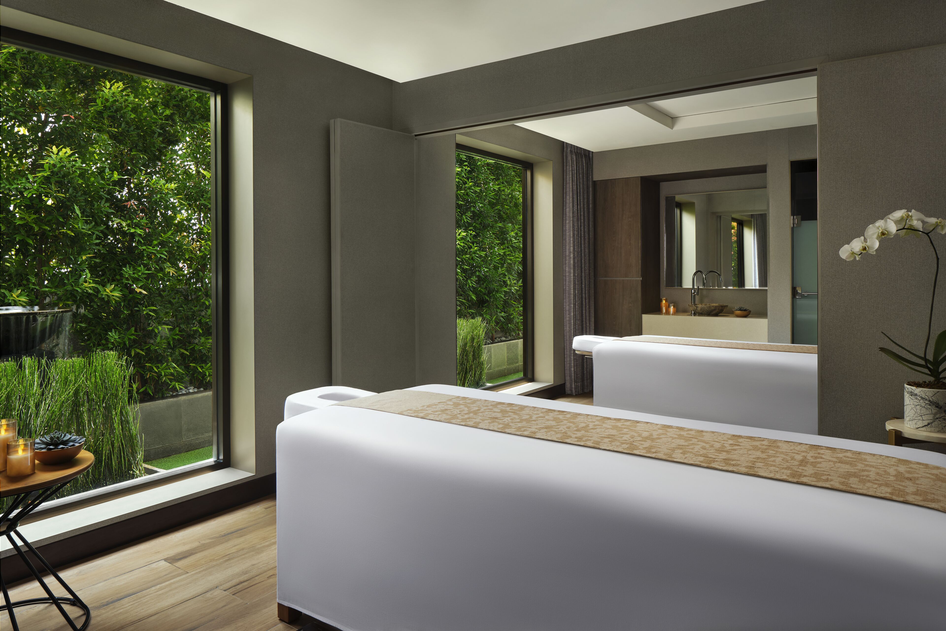 A modern spa room with a large window, a massage table, a small table with candles, and a potted plant.