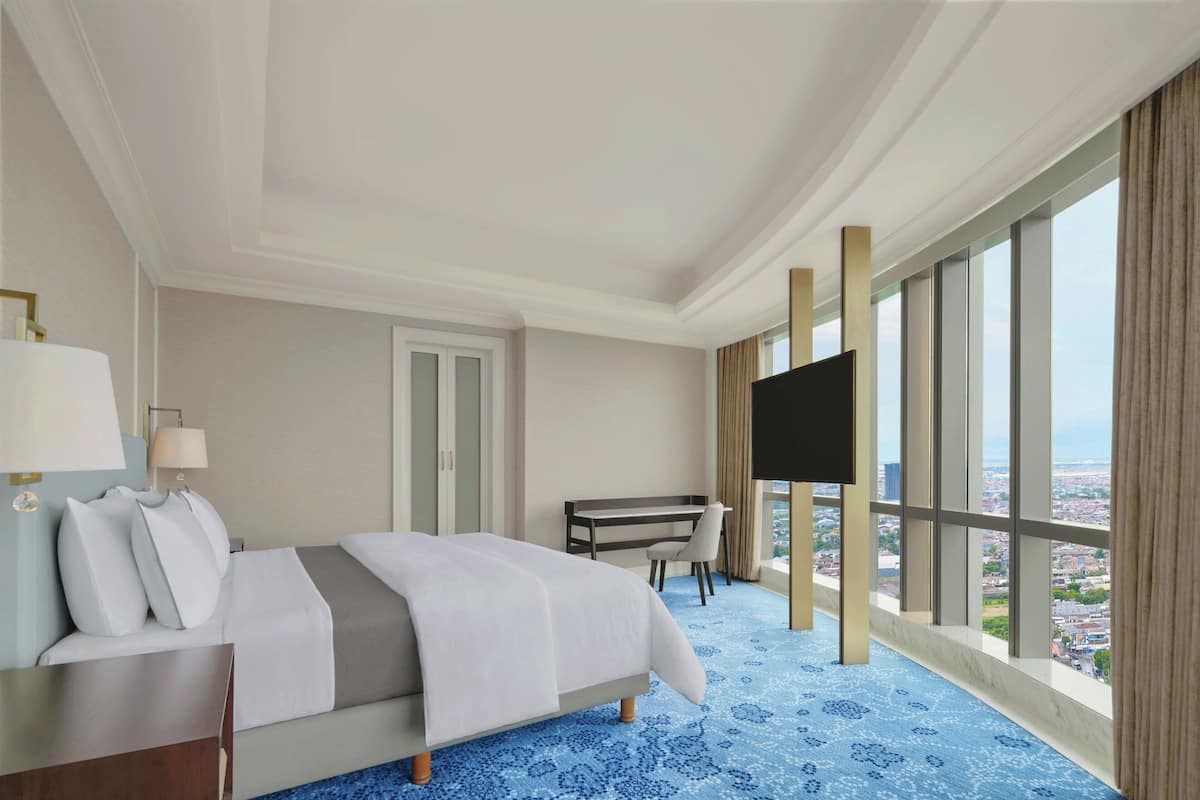 Habitación de hotel con cama, escritorio, silla, televisor y grandes ventanales con vistas a la ciudad.