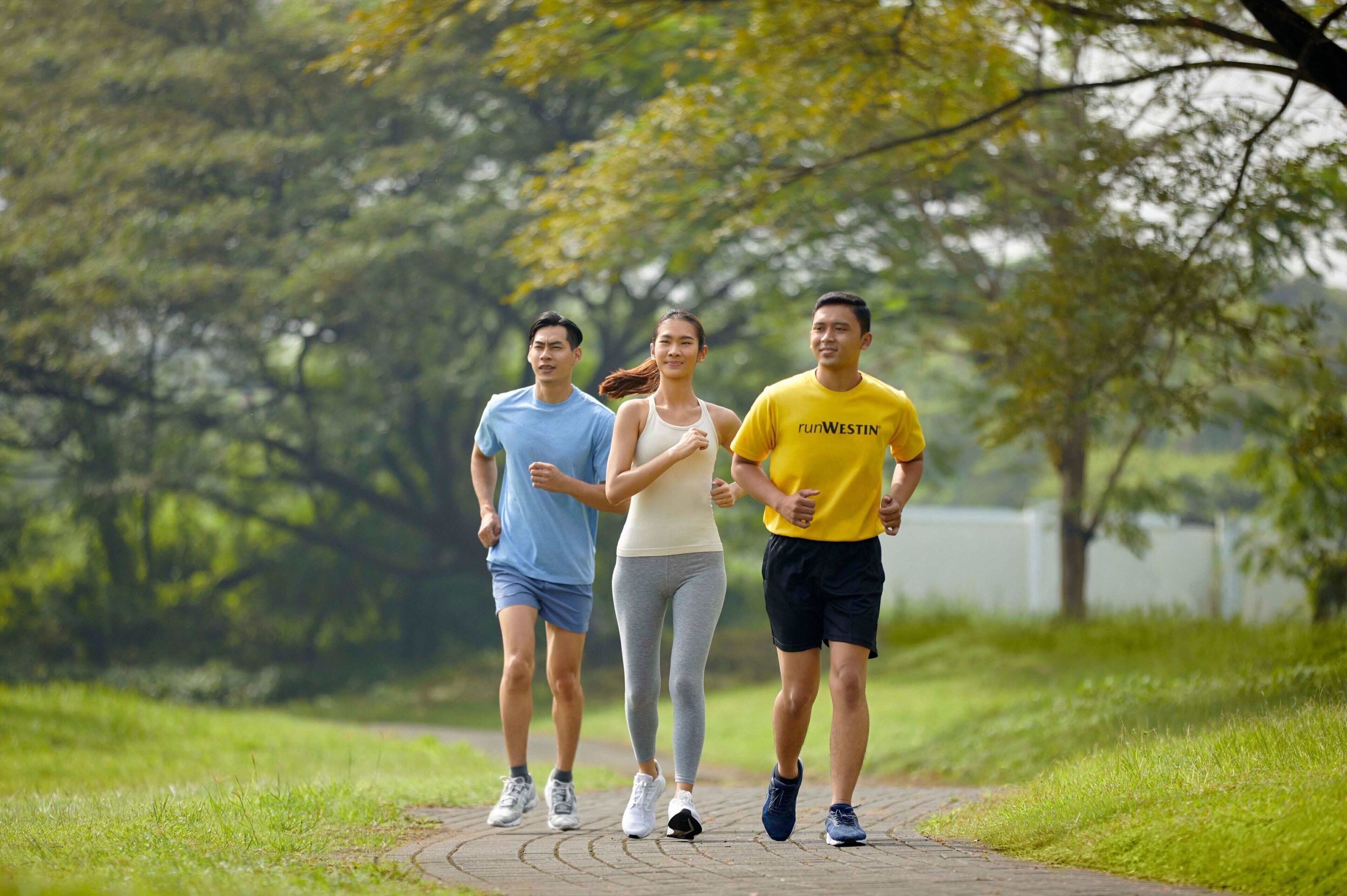 Tres personas trotando por un sendero pavimentado en un parque con árboles y césped al fondo.