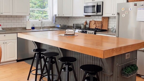 Une cuisine avec des meubles blancs, un îlot central en bois doté de tabourets, un four à micro-ondes et un réfrigérateur.