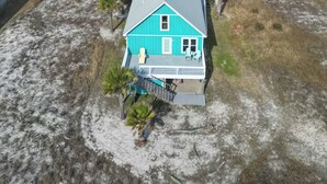 A single-story house with a blue exterior and a covered porch, surrounded by a dry landscape and a road leading to the ocean. - Beach “N” Bay Getaway Sleeps 9. Enjoy Beach Time,  Fishing, Kayaking. Steps Away (Gulf Shores)
