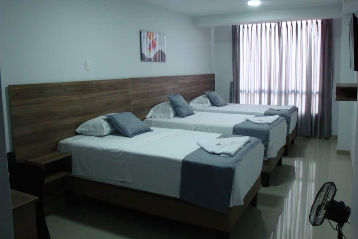 A hotel room with three beds, a wooden headboard, a flat-screen TV, and a standing fan.