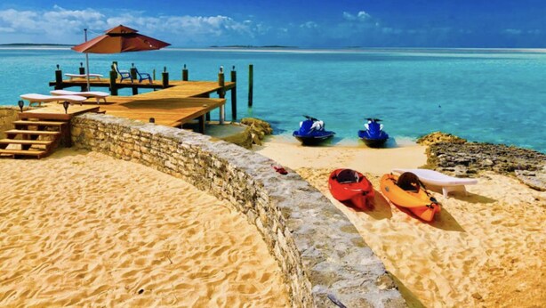A beach with a wooden pier, lounge chairs, and jet skis. - Amazing Beachfront home the calm side of the island. Swimming & kayaking (Exuma)