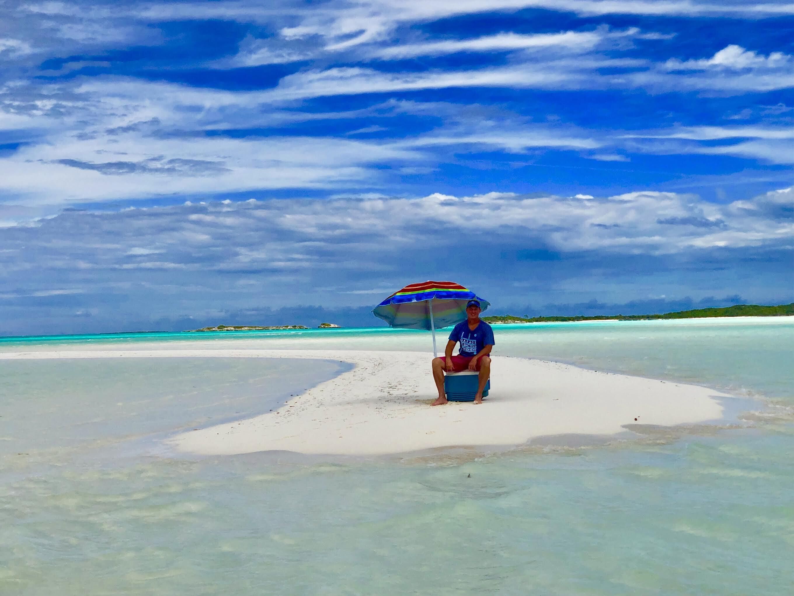 A person sitting under a colorful umbrella on a white sandy beach with clear turquoise water and a cloudy sky.