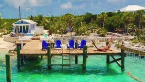 A dock with blue chairs and a small building, surrounded by clear turquoise water and palm trees. - Amazing Beachfront home the calm side of the island. Swimming & kayaking (Exuma)