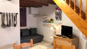 A cozy room with a sofa, a table with a patterned tablecloth, a TV, and a wooden staircase. - Homerez - House in Saint-Martin-de-Brômes (Saint-Martin-de-Brômes)