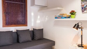 A grey sofa with cushions, a small table with a lamp, a window with a curtain, and a shelf with books and a plant. - Homerez - House in Saint-Martin-de-Brômes (Saint-Martin-de-Brômes)