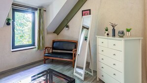 Una habitación con techo inclinado, una ventana con vistas a follaje, una silla de madera, un aparador blanco con cajones y un espejo apoyado en la pared.