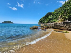 A beach with clear water, a sandy shore, and a rocky outcrop with greenery.