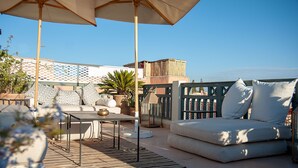 En udendørs terrasse med sofa, bord og parasol, med en mønstret væg og udsigt over byen.