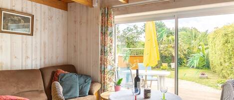 Une pièce chaleureuse avec un plafond en bois, un canapé, un fauteuil, une petite table avec des verres à vin et une vue sur un jardin, visible à travers une porte vitrée.