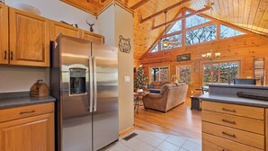 A cabin interior with a stainless steel refrigerator, wooden cabinets, a cozy living area with a fireplace, and a large window offering a view of trees. - Mountaintop Getaway with Gazeebo Deck Hot Tub & Fireplace! Long range views! (Bryson City)
