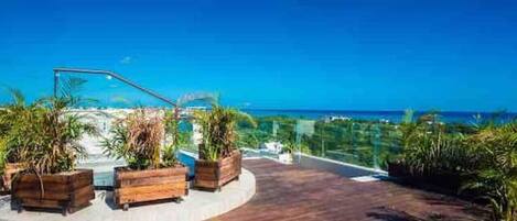 A rooftop terrace with wooden flooring, potted plants, and a view of the ocean.