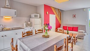Eine moderne Küche mit weißen Schränken, einer Mikrowelle und einer Kaffeemaschine. Ein Essbereich mit einem für zwei Personen gedecktem Tisch, umgeben von Holzstühlen. Im Hintergrund sind eine rote Wand und eine Treppe zu sehen.